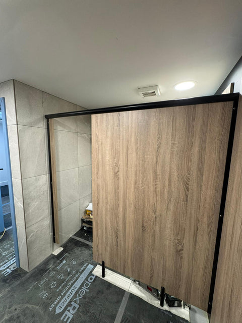 An inline Ascent Range toilet cubicle with a woodgrain door and black headrail, during installation