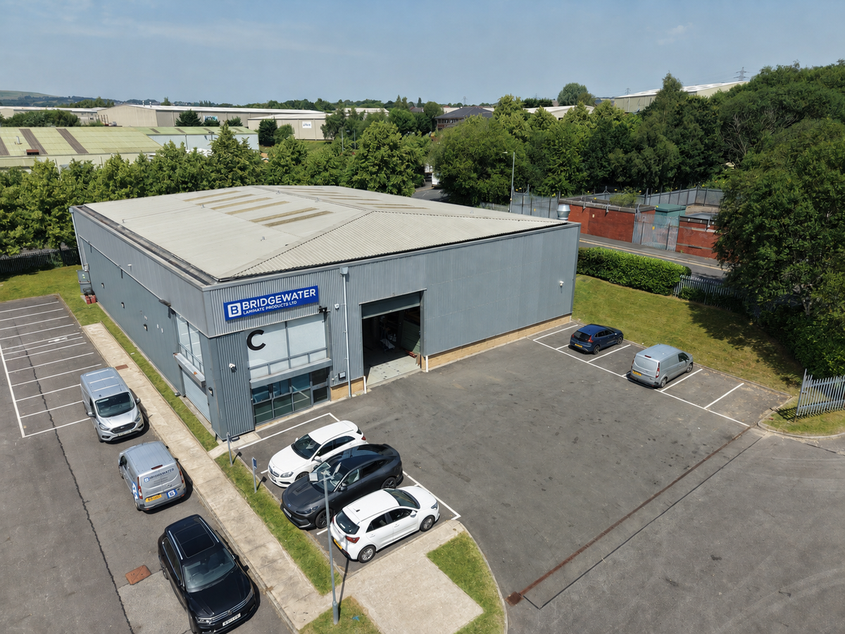 An aerial view of a gray industrial warehouse building with a blue and white Bridgwater sign, surrounded by a paved parking lot with several cars and green trees under a blue sky.