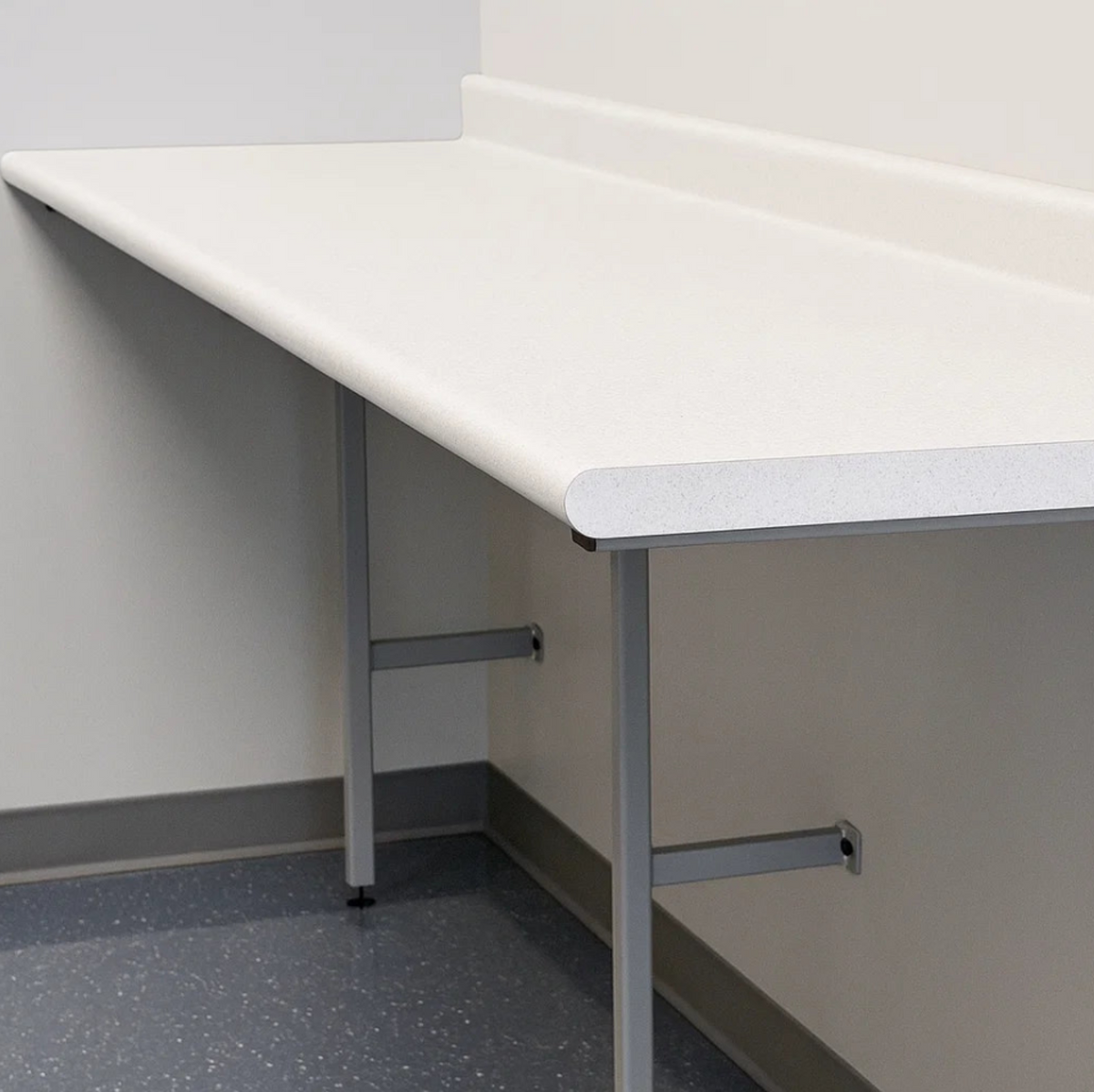 A white rectangular laboratory or work table with a thick apron edge and gray metal support legs, shown installed against a beige wall on a speckled gray floor.