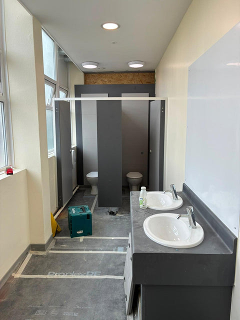 Grey Smart Range toilet cubicles being fitted in a washroom, beside a two-basin vanity unit