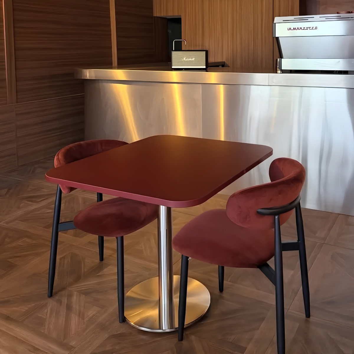 A modern square bar table with a reddish-brown top and a brushed metal pedestal base, accompanied by two matching velvet chairs in a warm-toned interior setting.